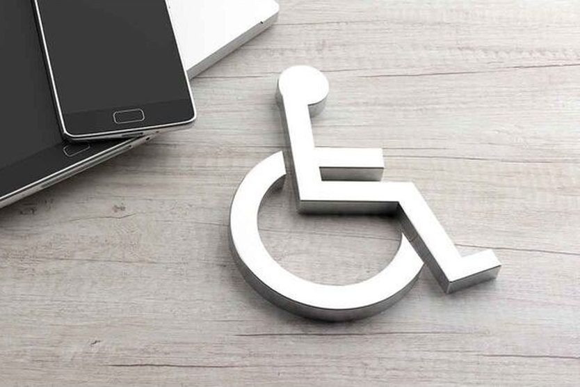 Weißes Rollstuhl-Symbol aus Metall liegt neben Laptop, Tablet und Smartphone auf einem Holztisch