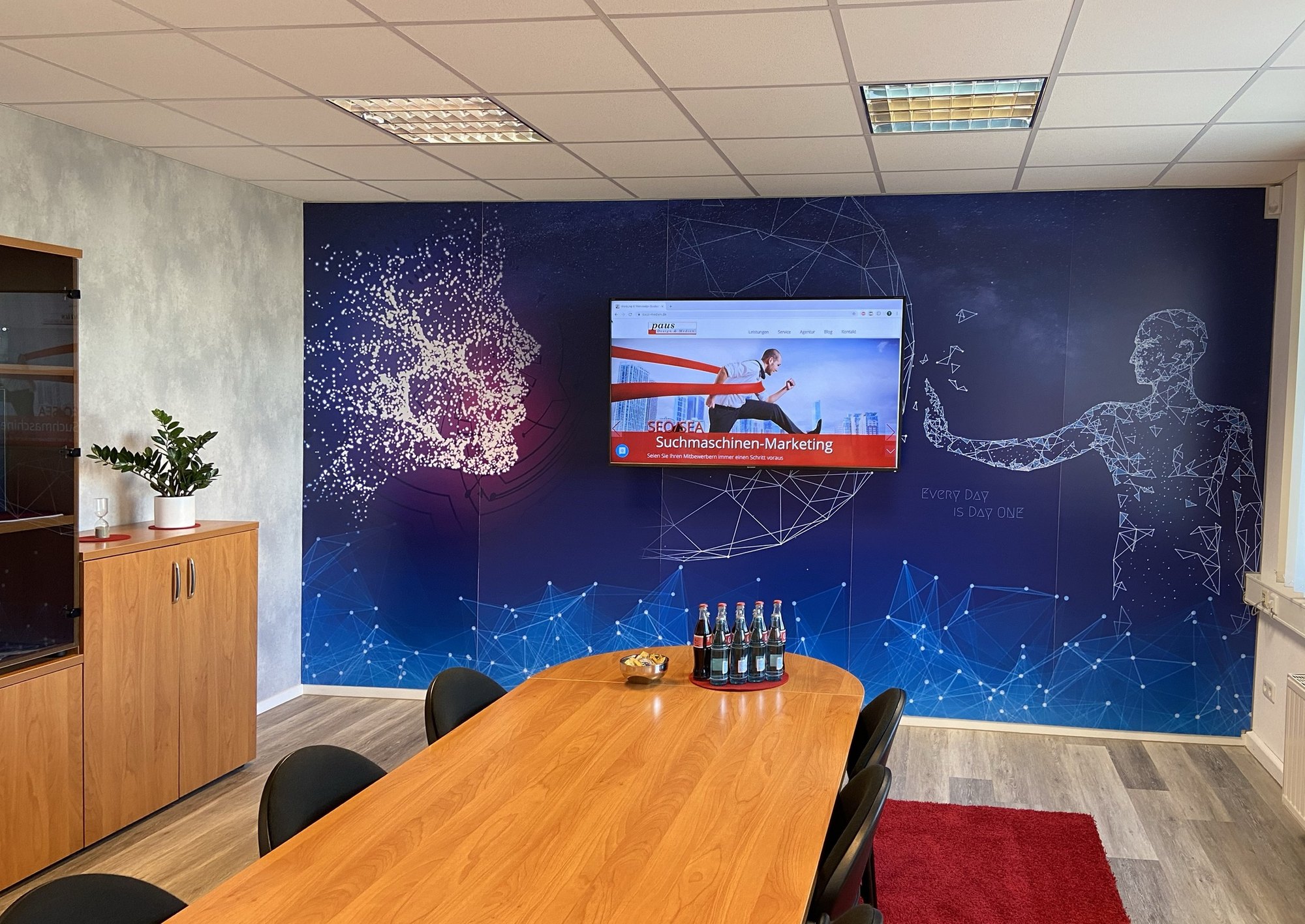 Besprechungsraum der Agentur: langer Holztisch vor einer dunkelblauen Wand mit Sternbild-Motiv und einem Bildschirm