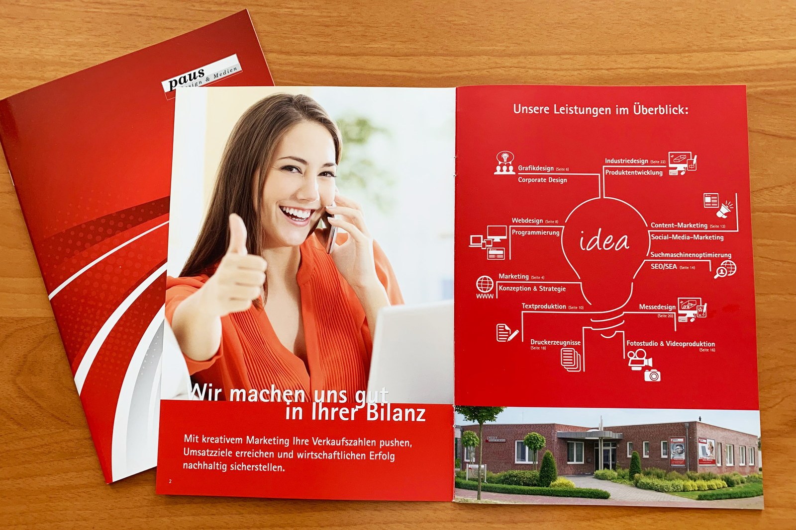 Aufgeschlagene Imagebroschüre von Paus Design & Medien auf einem Holztisch: links eine telefonierende Frau mit erhobenem Daumen, rechts die Leistungen im Überblick als Glühbirne, dahinter das rote Titelblatt
