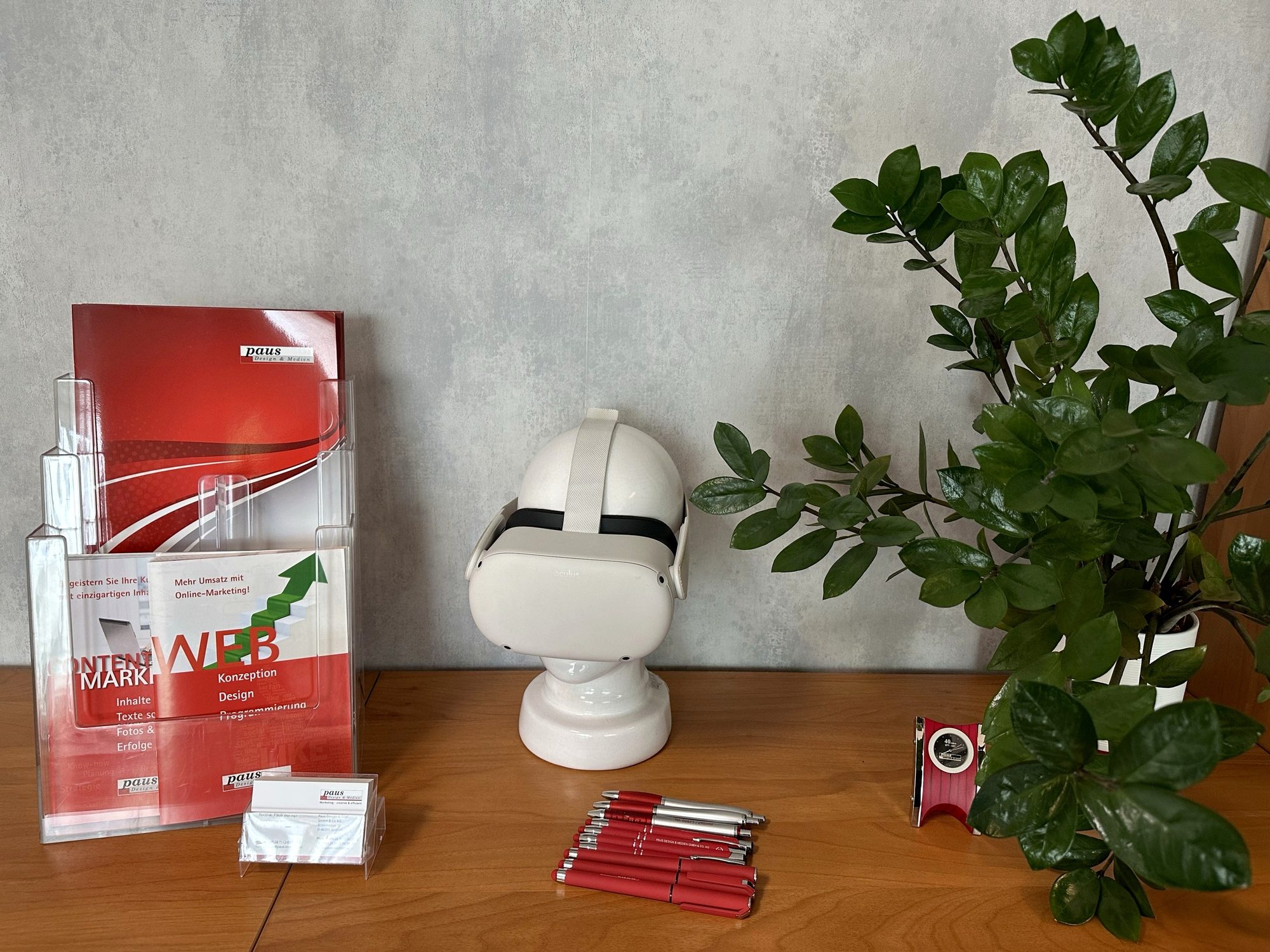 Prospektständer mit Imagemappe und Flyern von Paus Design & Medien, Visitenkarten und rote Kugelschreiber, in der Mitte eine VR-Brille auf einer Kopfbüste, rechts eine Zimmerpflanze