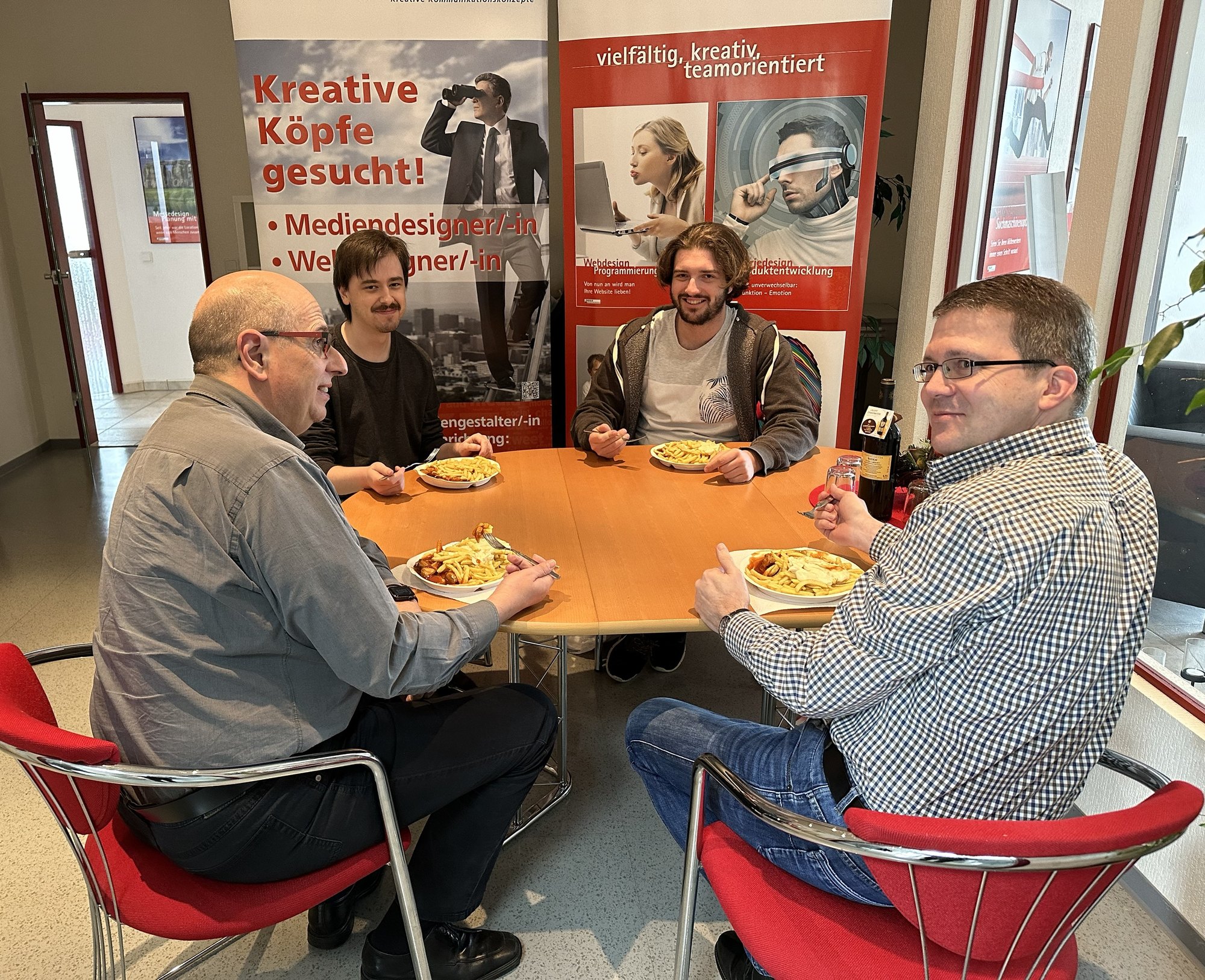 Vier Kollegen essen gemeinsam am runden Tisch in der Agentur, im Hintergrund das Roll-up „Kreative Köpfe gesucht!“