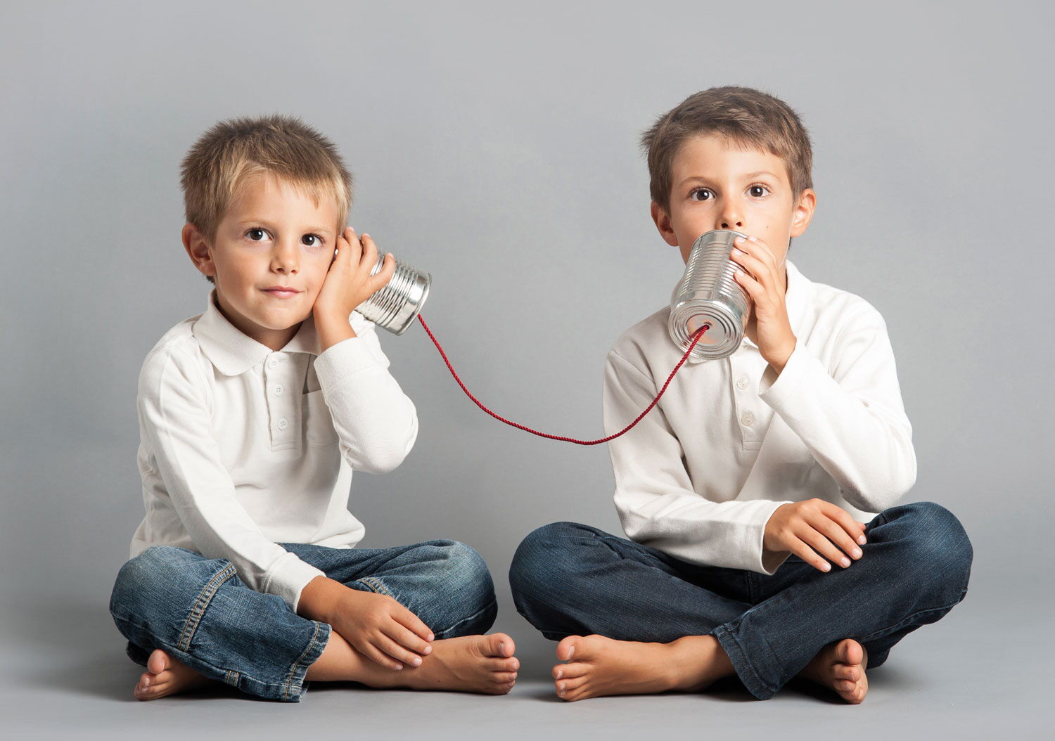 Zwei Jungen sitzen auf dem Boden und telefonieren über zwei Konservendosen, die mit einer roten Schnur verbunden sind