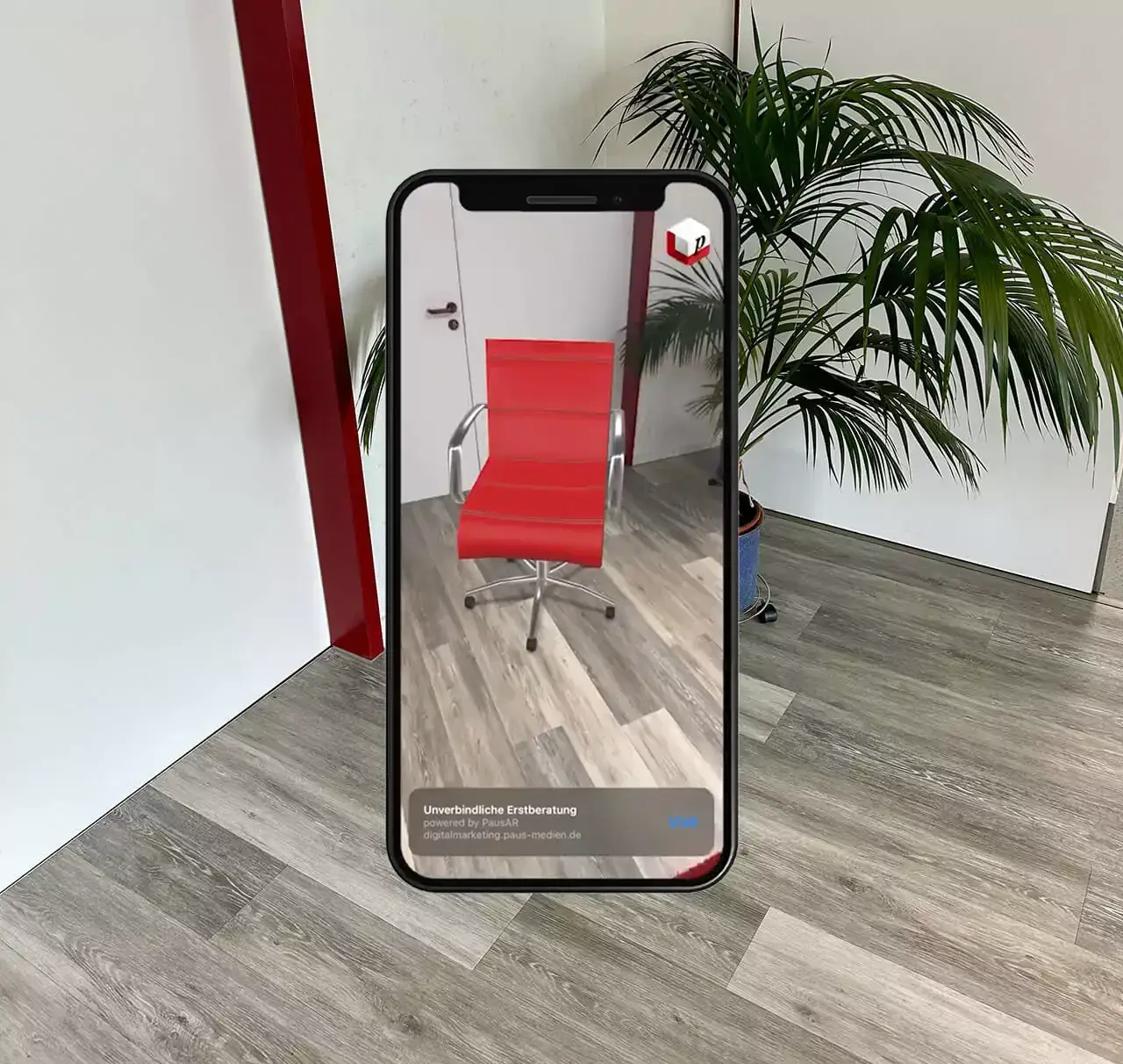 Smartphone vor einer weißen Wand mit Zimmerpalme, auf dem Display steht ein roter Bürostuhl per Augmented Reality auf dem Holzboden, darunter ein Banner mit Kontaktknopf