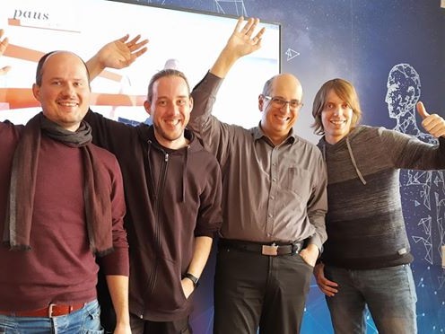 Das IT-Team von Paus Design und Medien mit erhobenen Händen vor einer Leinwand mit dem Paus-Logo, von links: Marinko, Marc, Nicolai Paus und Holger